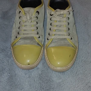 Lanvin sneakers med gul tå i mocka - Snygga sneakers från Lanvin med beige mocka och kontrasterande gul lackad tå. Skorna har rund tå, platt sula och snörning med metallöglor. Insidan är svart och sulan är ljusbeige. Perfekt för dig som gillar stilrena detaljer med en twist.