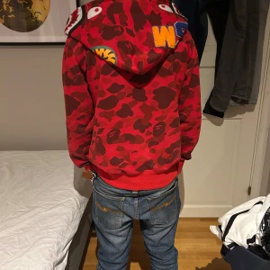 Röd Bape hoodie camouflage  - Säljer en röd camo hoodie från Bape med ikonisk hajdesign på huvan, broderade bokstäver och tigerpatch. Pris kan diskuteras vid snabb affär. Hör av dig vid intresse!😃