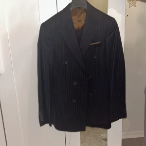 Svart randig dubbelknäppt kostymkavaj - Snygg svart dubbelknäppt kostymkavaj med tunna vita ränder och klassisk passform. Kavajen har bruna detaljer på insidan och vid bröstfickan, samt flera fickor och knappar framtill. Perfekt för dig som gillar stilrena och tidlösa plagg. Använd en gång 