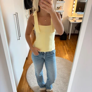 Gul topp med rosett från Zara - Säljer en ljusgul topp från Zara i storlek XS. Toppen har breda axelband med snygga rosetter på axlarna och en fyrkantig urringning. Materialet är slätt och känns som polyester. Perfekt för dig som gillar stilrena och söta detaljer.