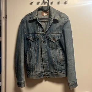 Klassisk jeansjacka från Levi's - Säljer en klassisk blå jeansjacka från Levi's med snygga bruna knappar och två bröstfickor med lock. Jackan har lång ärm, krage och justerbara detaljer vid midjan. Perfekt för dig som gillar tidlös streetstil och vill ha en jacka som funkar året runt.
