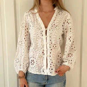 Vit broderad blus med knappar - Superfin vit blus i bomull med broderade blommönster och genombrutna detaljer. Blusen har v-ringning, knappar framtill och långa puffiga ärmar med resår vid handlederna. Perfekt för en fräsch och somrig look.