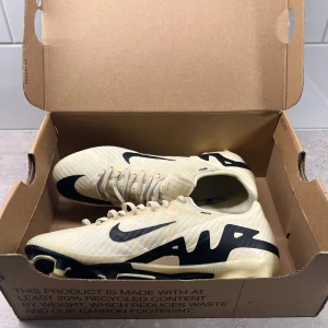 Nike Air Zoom  - Säljer ett par Nike Air Zoom fotbollsskor i beige med svarta detaljer och stor Nike-logga på sidan. Skorna har snörning och en låg profil med platt sula och rund tå. Perfekta för dig som vill ha snygga och funktionella skor på planen.