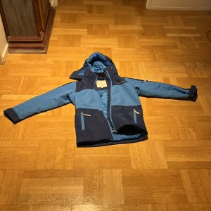 Blå vindjacka från Nordbjørn, strl 140 - Säljer en blå vindjacka från Nordbjørn i storlek 140. Jackan har två nyanser av blått, huva och dragkedjor framtill samt på fickorna. Perfekt för blåsiga dagar och har en sportig look med praktiska detaljer.