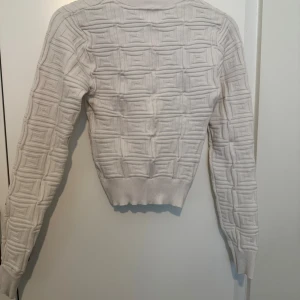 Vit stickad tröja med 3D-mönster - Säljer en vit stickad tröja med unikt 3D-mönster i geometriska rutor. Tröjan har lång ärm, ribbade muddar och en tajtare passform i midjan. Perfekt för dig som vill ha något stilrent men ändå annorlunda.
