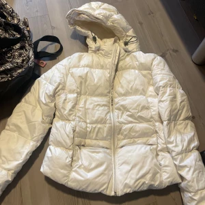 Vit pufferjacka  - Säljer en vit pufferjacka från Nike med huva och dragkedja framtill. Jackan är quiltad och har en clean look med breda kanaler och dragsko i huvan. Perfekt för kalla dagar när du vill hålla stilen enkel men ändå varm.