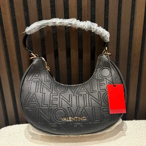 Svart axelväska från Valentino - Köpte väskan för 1495kr säljer den för endast 500kr den är Snygg svart axelväska från Valentino med präglad logotyp över hela väskan och guldfärgade detaljer. Väskan har en rundad form, dragkedja upptill och ett brett handtag. Materialet är skinn med struktur och väskan har en röd tagg från märket. Väskanr helt ny med prislapp på aldrig använd . Medföljer två st band 