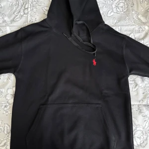 Svart hoodie från Polo Ralph Lauren - Svart hoodie från Polo Ralph Lauren med klassisk röd broderad logga på bröstet. Tröjan har huva med snörning och en stor magficka framtill. Tillverkad i mjuk bomull, perfekt för chill dagar eller när du vill ha en clean streetstyle.