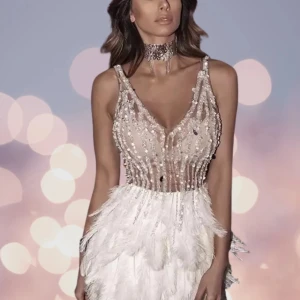 Bella Barnett - Superfin kortklänning med glittrig överdel i nude och silver, täckt av paljetter och pärlor. Kjolen är dekorerad med vita fjädrar som ger en lyxig känsla. Djup v-ringning och smala axelband. Perfekt för dig som vill sticka ut på bal eller fest! Ubrukt. Nypris var 1300.  