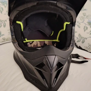 NOLAN Svart hjälm med visir för moped, cross och MC - Säljer en svart crosshjälm från Noxon med cool, kantig design och skärm framtill. Invändigt har hjälmen meshfoder med neongula detaljer och camouflagemönster. Justerbart hakspänne och bra ventilation. Perfekt för dig som vill ha en snygg och funktionell hjälm till cross eller liknande.