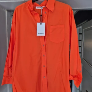 Oversized orange skjorta från Co'couture - Snygg oversized skjorta i stark orange färg från Co'couture. Skjortan är tillverkad i 100% bomull, har klassisk krage, knappstängning framtill och en bröstficka. Perfekt för dig som gillar att sticka ut med färg och vill ha en chill, loose passform. Helt ny.