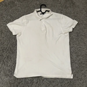 Vit klassisk pikétröja med krage - Vit pikétröja med klassisk krage och korta ärmar. Tröjan har tre knappar framtill och är tillverkad i mjuk bomull. Enkel och stilren design som funkar till många olika looks. Perfekt för dig som gillar clean och tidlös stil.