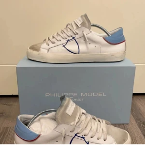 Philippe Model vita sneakers med blå detalj - Snygga vita Philippe Model sneakers med blå och röda detaljer vid hälen och beige mocka framtill. Skorna har klassisk snörning, platt sula och en blå sköldlogga på sidan. Perfekta för dig som gillar stilrena men unika sneakers. Ganska nya änvnät dom 2 gånger köpte dom ny pris 3500 så passa på, några funderingar skriv bara till mig!
