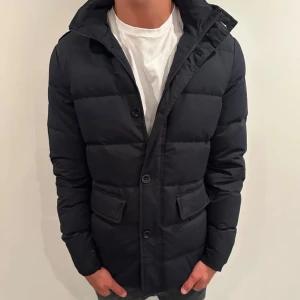 Mörkblå pufferjacka med knappar - Hej, säljer nu denna riktigt snygga jackan som liknar en herno jacka! Den är i storlek M och är perfekt nu till hösten! Skicket är 9/10 vid minsta lilla fundering är det bara att höra av sig, finns små fläckar vid ärmarna men de syns inte vid användning, kontakta för bilder!