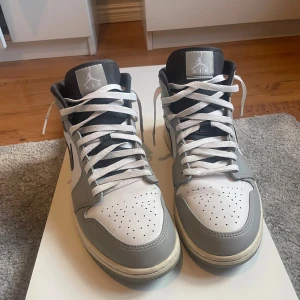Nike Air Jordan 1 Mid Grå/Vit US 11.5 - Säljer ett par Nike Air Jordan 1 Mid i grått och vitt med svarta detaljer. Klassisk high-top siluett med svart swoosh, perforerad tå och vita skosnören. Ovandel i skinn och syntet, ikonisk Jordan-logga på sidan. Perfekta för dig som vill sticka ut med stil.