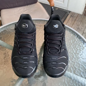 Nike Air Max Plus Tn helsvarta sneakers - Säljer ett par helsvarta Nike Air Max Plus Tn sneakers med klassisk vågig design och synliga Air Max-bubblor i sulan. Skorna har mesh- och syntetmaterial, snörning och diskreta Tn-detaljer på hälen. Perfekta för dig som gillar streetwear och sportig stil.