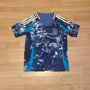 Ajax blå bortatröja Adidas - Ajax bortatröja i blått med abstrakt mönster och ljusblå detaljer. Klassiska Adidas-ränder på axlarna, klubbmärke på bröstet och tryck med namn och nummer (Godts 39) på ryggen. Tillverkad i lätt och ventilerande material, perfekt för fotbollsträning eller match.