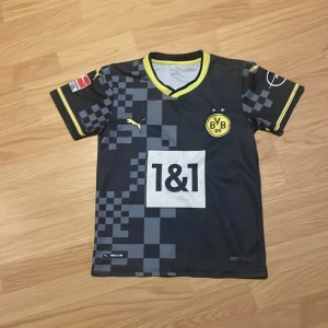 Borussia Dortmund Bellingham tröja - Borussia Dortmund matchtröja med Bellingham 22 på ryggen. Svart med gula detaljer och rutigt mönster på ärmarna. Puma-logga, Bundesliga-märke och sponsortryck. Tillverkad i lätt polyester med V-ringad krage och korta ärmar.