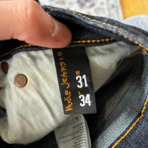 Nudie Jeans High Kai mörkblå jeans - Mörkblå jeans från Nudie Jeans, modell High Kai. Slimmad passform med klassiska fem fickor och snygga kontrastsömmar. Tillverkade i 98% ekologisk bomull och 2% elastan för extra stretch och komfort. Perfekta för dig som gillar en stilren och modern look.
