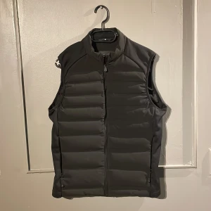 Svart dunväst från Sail Racing - Svart dunväst från Sail Racing med quiltad design och hög krage. Västen har dragkedja framtill och två sidofickor med dragkedja. Perfekt för lager-på-lager och stilren look. Passar både till hoodie och t-shirt.