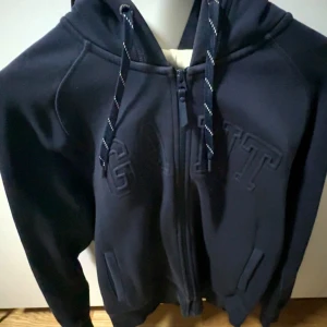 Svart hoodie med dragkedja från GANT - Säljer en svart hoodie från GANT i storlek L. Tröjan har dragkedja framtill, broderad GANT-logga på bröstet, snörning i huvan och två fickor. Perfekt för dig som gillar en klassisk och stilren look.