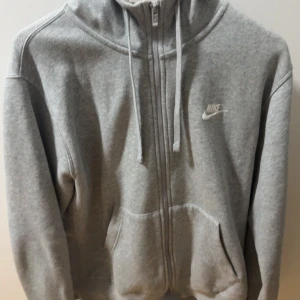 Grå hoodie från Nike med dragkedja - Grå hoodie från Nike med hel dragkedja, huva med snörning och två stora fickor framtill. Mjuk fleece på insidan för extra komfort. Perfekt för chill dagar eller när du vill ha en sportig look.