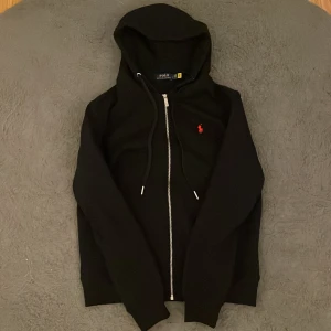 Svart hoodie från Polo Ralph Lauren - Svart hoodie från Polo Ralph Lauren med dragkedja och huva. Klassisk röd broderad logga på bröstet och snörning i huvan. Tillverkad i mjuk bomullsmix, perfekt för chill dagar. Snygg och enkel att matcha med allt.