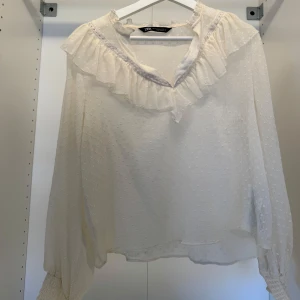 Vit blus med volang från Zara - Supersöt vit blus från Zara med volangdetalj runt halsen och diskret prickigt mönster. Blusen har långärmade ärmar med smock vid ärmslut och en lätt transparent känsla. Perfekt för dig som gillar romantisk och feminin stil.