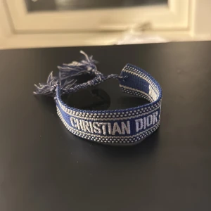 Blå Christian Dior J'Adior armband - Snyggt blått armband från Christian Dior med broderad text 'CHRISTIAN DIOR' och 'DIOR J'ADIOR' i vitt. Armbandet har flätade snören och tofsar i blått och vitt som ger en cool och trendig vibe. Perfekt accessoar för att lyfta din outfit.