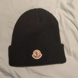 Svart Moncler mössa - Ribbstickad svart mössa från Moncler med klassisk uppvikt kant och broderad logotyp framtill. Tillverkad i mjuk ull som håller dig varm under kyliga dagar. Perfekt accessoar för en clean och stilren look.