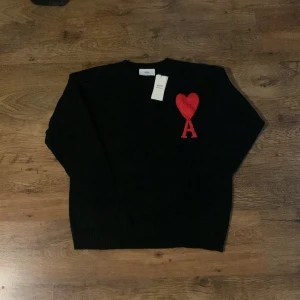 Svart stickad tröja från Ami Paris - Svart stickad tröja från Ami Paris med röd broderad hjärtlogga och bokstaven A på bröstet. Klassisk rund halsringning och långa ärmar. Perfekt för dig som gillar stilrena plagg med en twist.