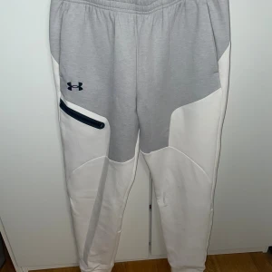 Gråvita mjukisbyxor från Under Armour - Snygga mjukisbyxor från Under Armour i grått och vitt med svart logga på vänster lår. Byxorna har elastisk midja, en dragkedjeficka på sidan och coola paneldetaljer. Perfekta för chill eller träning, riktigt sköna och stilrena. Är i ett fint skick, men säljer då de inte används längre, hör av er vid frågor💕
