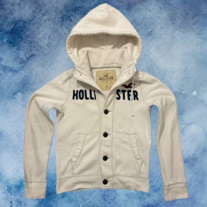 Vit Vintage Hollister Cardigan/Kofta Med Knappar - Väldigt sällsynt och vintage Hollister kofta/caridgan. Tröjan har stora knappar framtill, dragsko i huvan och två fickor. (Högra armen har 2 jätte små fläckar som går att tvättas bort, dessutom ett pyttelitet hål som inte alls märks då hålet göms av koftans färg! defekterna ställer inte till med problem då vintage koftorna har en mer distressed/använd look) Längd 63cm, bredd 52-53cm. Storlek M