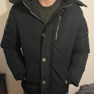 Svart pufferjacka från Moose Knuckles - Säljer en svart pufferjacka från Moose Knuckles med huva, silverfärgade dragkedjor och knappar. Jackan har ett snyggt metallmärke på ärmen och broderad patch på insidan. Flera fickor med dragkedja och detaljer som ger jackan en cool vibe. Ull på dragkedjan och den har ett litet hål i vendetta fickan.
