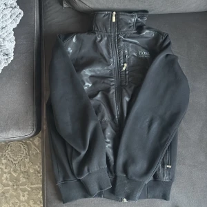 Svart vindjacka från Hugo Boss - Svart vindjacka från Hugo Boss med diskret camouflagemönster på framsidan. Jackan har dragkedja, huva och ribbade muddar. Märkeslogga broderad på bröstet och praktisk bröstficka med dragkedja. Perfekt för dig som vill ha en stilren och sportig look.