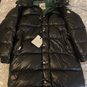 Svart Moncler pufferjacka med huva (lång) - Svart pufferjacka från Moncler med glansig finish, huva och dragkedjor på sidorna. Jackan har klassisk Moncler-logga på ärmen, tryckknappar med logotyp och grönt innerfoder. Perfekt för kalla dagar och riktigt snygg streetstil.