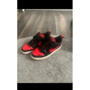 Nike Dunk Low svart/röd sneakers - Snygga Nike Dunk Low sneakers i svart och rött med klassisk vit sula. Skorna har snörning och är tillverkade i skinn och syntet. Perfekta för dig som vill sticka ut med en ikonisk streetstyle-look.