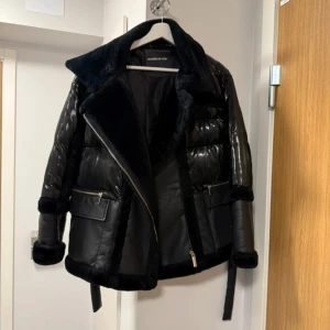 Svart pufferjacka från Manière De Voir - Snygg svart pufferjacka från Manière De Voir med bred krage och detaljer i fuskpäls. Jackan har glansiga paneler, dragkedja framtill och fickor med zip. Bältet i midjan ger extra edge och jackan har en cool, oversized vibe.