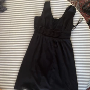 Svart kort klänning Vero Moda S - Svart klänning från Vero Moda i storlek S. Modellen är kort med breda axelband och har ett snyggt veckat midjeband. Omlott-detalj med spets vid urringningen ger en extra touch. Materialet känns lätt och har en fin lyster.