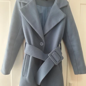 Ljusblå kappa med bälte och stora slag - Snygg ljusblå kappa med breda slag, markerat midjebälte och knäppning framtill. Jackan har en klassisk siluett och är helfodrad. Perfekt för dig som vill ha en stilren och trendig look under kallare dagar.
