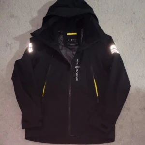Svart Sail Racing windbreaker - Svart windbreaker från Sail Racing med huva i storlek S för män, helt oanvänd. Perfekt för blåsiga dagar.