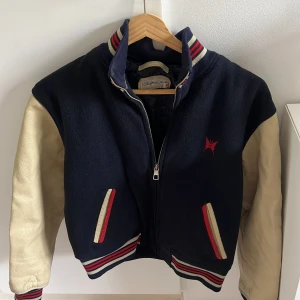 Clubjacka från Chevignon Girl M - Mörkblå clubjacka med krämvita skinnärmar och broderad röd fjäril och texten 'Manhattan Angel' på ryggen. Jackan har randiga muddar i rött, vitt och blått, dragkedja framtill och coola detaljer vid fickorna. Med ärmar av äkta läder