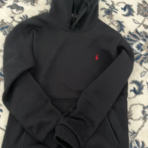 Svart hoodie från Polo Ralph Lauren - Svart hoodie från Polo Ralph Lauren med klassisk röd broderad logga på bröstet. Tröjan har huva med snörning och en stor magficka. Perfekt för dig som gillar stilren och enkel streetwear.