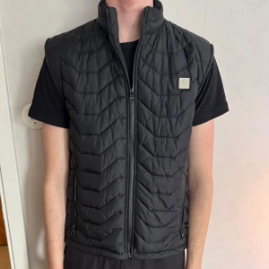 Svart ä dunväst  - Svart dunväst från SAMS BLACK med quiltat mönster i sicksack, hög krage och dragkedja framtill. Västen har två fickor med dragkedja och en liten logga på bröstet. (Katt finns i hemmet) nypris 500kr (även lite nopprig på insidan men de går att ta bort) 