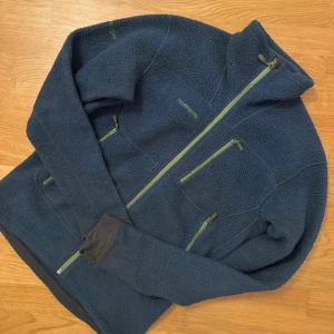 Blå fleecejacka från Norrøna Trollveggen - Mysig mörkblå fleecejacka från Norrøna Trollveggen med gröna dragkedjor och broderad logga. Jackan har hög krage, långa ärmar med tumhål och flera praktiska fickor med dragkedja. Perfekt för kalla dagar och outdoor-häng.