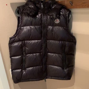 Monclerväst svart - Snygg svart dunväst från Moncler med glansig finish, avtagbar huva och två dragkedjefickor framtill. Västen har klassisk Moncler-logga på bröstet och quiltad design. Perfekt för lager-på-lager under höst och vinter.