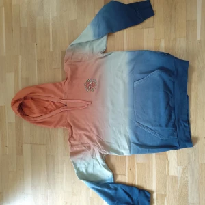 Vans hoodie "hi on peace" - Säljer en hoodie med cool färgövergång från orange till blå och ett broderat märke framtill. Baksidan har ett stort tryck med texten 'HI ON PEACE' och regnbågsmotiv. Tröjan har huva med snörning och stor magficka. Perfekt för dig som gillar unika plagg!