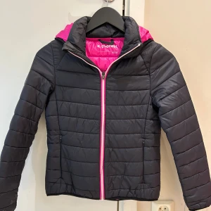 Barn Svart pufferjacka från Everest - Snygg svart pufferjacka från Everest med rosa foder och dragkedja. Jackan har huva, quiltad design och långa ärmar. Perfekt för dig som vill ha en varm och stilren jacka med lite färgklick. Passar dig som gillar sportig och modern stil.