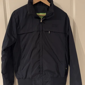 Svart vindjacka från Hugo Boss - Snygg svart vindjacka från Hugo Boss med klassisk design. Jackan har hög krage, dragkedja framtill och diskret logga på bröstet. Tillverkad i lätt polyester och har ribbade muddar i midja och ärmslut. Perfekt för dig som gillar stilren och sportig look. Pris ej fast kom med frågor🤗