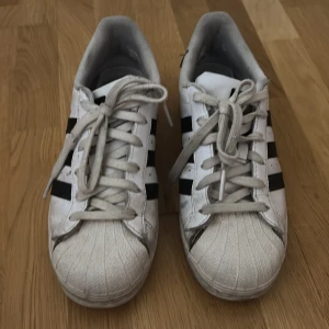 Adidas Superstar vita sneakers - Klassiska Adidas Superstar sneakers i vitt med svarta ränder och logga på hälen. Skorna har snörning, rund tå och platt sula. Ovandelen är i skinn och insidan är fodrad med grå textil. Perfekt för dig som gillar retrostil och streetwear.